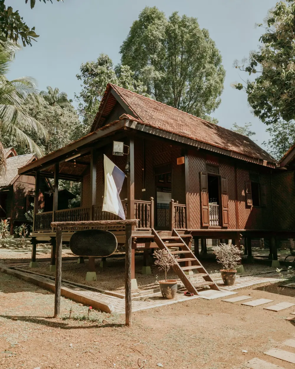 A traditional Malay timber house raised on stilts, with a shingled roof, front veranda and steps, surrounded by trees